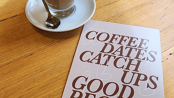 Almond latte with a heart drawn with the milk, it's in a glass little cup, and there's a small spoon on its side, over a wooden table with a menu that reads coffee dates, catch ups good people with a light brown background, brown letters and some white doodles.