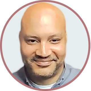 Smiling man with a shaved head in a blue shirt, centered in a circular frame with a light gray background. Mood appears friendly and calm.