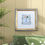 Thumbnail: Framed Hebrew Home Blessing with a Golden House artwork, in a lounge, hung above a couch and a pot plant.