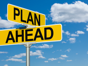 clear blue sky with scattered white clouds in the background. In the foreground, there is a bold yellow road sign mounted on a metal pole. The sign is split into two panels: the top panel reads “PLAN” and the bottom panel reads “AHEAD,” both in large black capital letters.