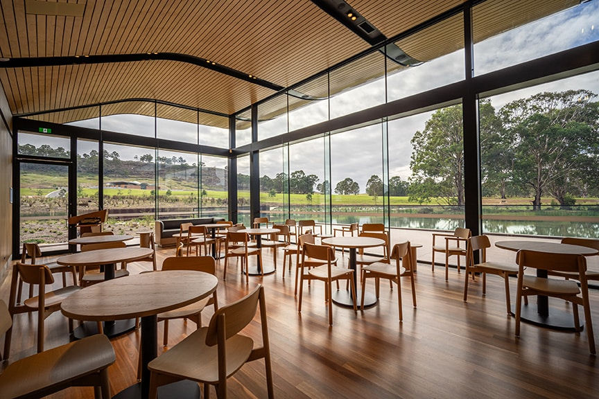 Modern cafe with wooden tables and chairs, large glass windows showing a scenic view of trees and a pond. Bright and serene.