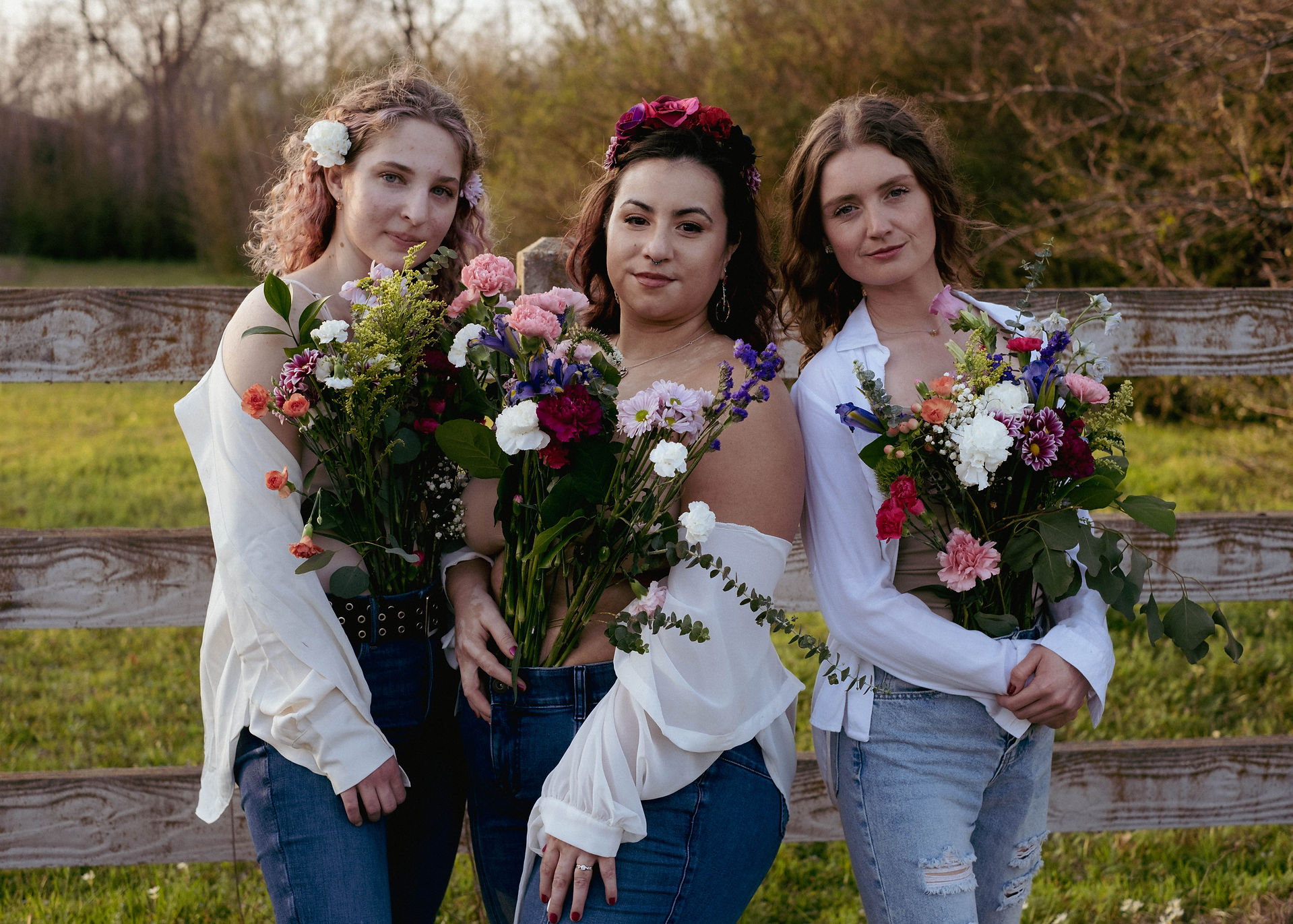 Blond woman with curly hair, woman with vitiligo, and brown haired woman standing together holding bouquets of flowers.