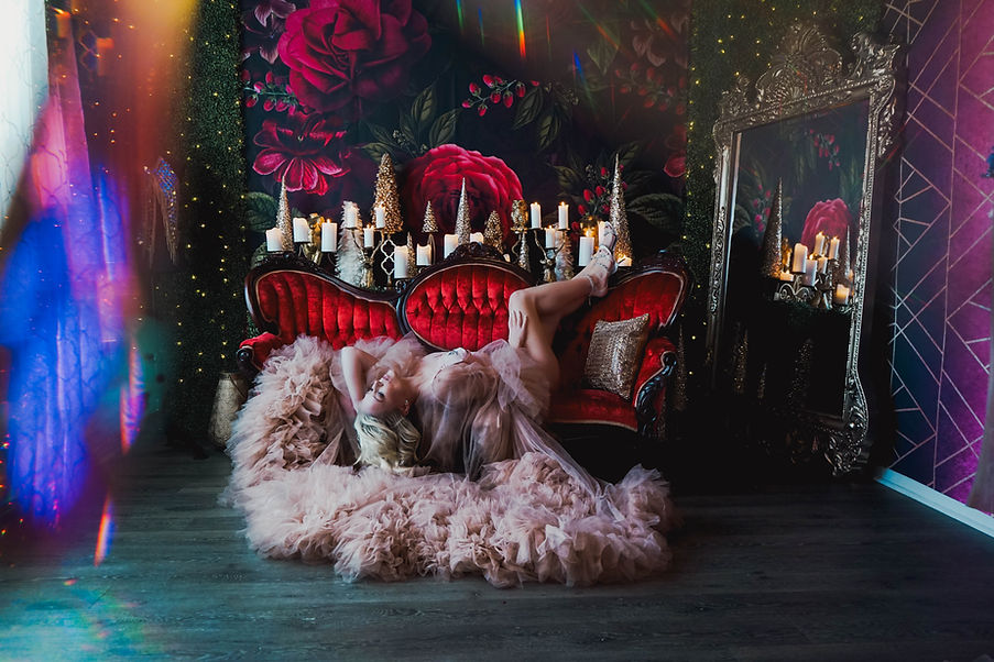 A thin middle-aged female wearing a pink tulle robe laying on a red Victorian couch in a desirable pose.
