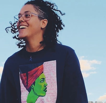 Woman wearing glasses, and a long sleeved tee in navy blue, with an image drawn by Enivid by B Genette of a green-skinned woman wearing a red headwrap with locs/braids and a pink background.