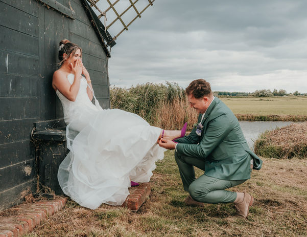 Groom kneels to remove the bride’s garter outdoors near a rustic building at a Cyprus wedding.