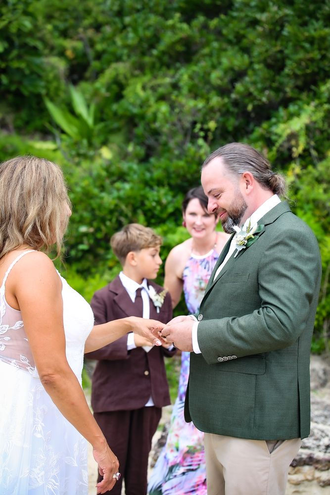 Bride and groom exchange rings outdoors, surrounded by greenery. A young boy and celebrant in floral dress look on, creating a joyful, calm mood.