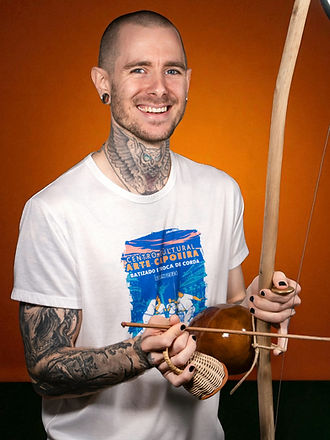 Instrutor Modelo lächelt und spielt Berimbau, trägt ein Capoeira Arte T-Shirt vor orangem Hintergrund