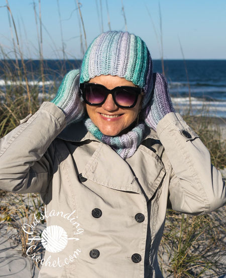 A woman wearing a coordinated Ryft crochet set in shades of green and purple. She is pictured from the chest up, sporting a chunky ribbed beanie pulled low, a thick matching cowl, and fingerless gloves. The Aran weight cotton yarn shows a seamless transition between colors, following the vertical texture of the ribs.