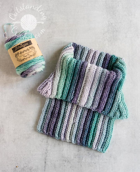 A flat lay of a unisex cowl neck warmer. The multicolored yarn creates a seamless transition between vertical stripes, highlighting the thick, vertical ribbing texture of the Aran weight cotton yarn in green and purple shades.