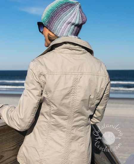 A woman wearing a coordinated Ryft crochet set in shades of green and purple. She is pictured from the chest up, sporting a chunky ribbed beanie pulled low, a thick matching cowl, and fingerless gloves. The Aran weight cotton yarn shows a seamless transition between colors, following the vertical texture of the ribs.