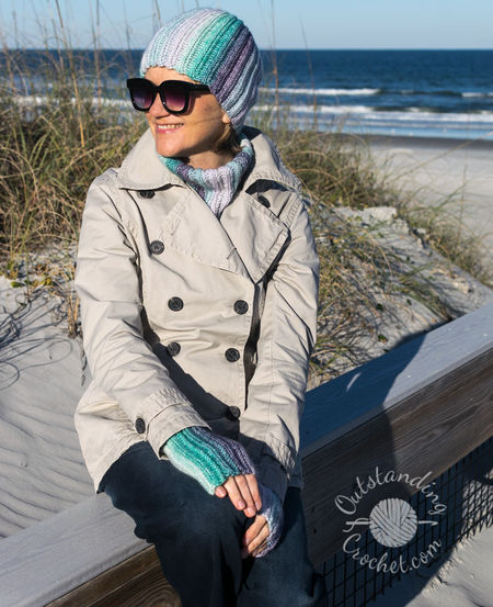 A woman wearing a coordinated Ryft crochet set in shades of green and purple. She is pictured from the chest up, sporting a chunky ribbed beanie pulled low, a thick matching cowl, and fingerless gloves. The Aran weight cotton yarn shows a seamless transition between colors, following the vertical texture of the ribs.