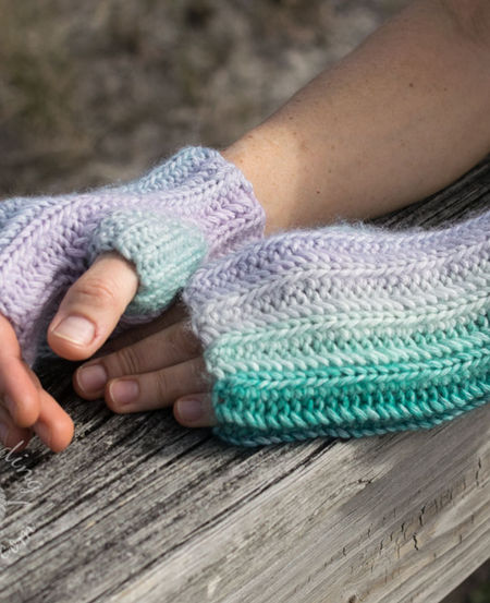 Pictures shows hands in Ryft unisex fingerless gloves. The multicolored yarn creates a seamless transition between vertical stripes, highlighting the thick, vertical ribbing texture of the Aran weight cotton yarn in green and purple shades.