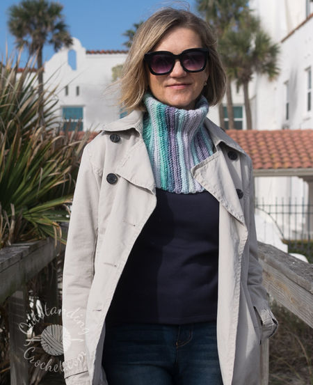 A woman wearing Ryft Crochet Neck Warmer in shades of green and purple. She is pictured from the chest up, sporting a chunky cowl. The Aran weight cotton yarn shows a seamless transition between colors, following the vertical texture of the ribs.