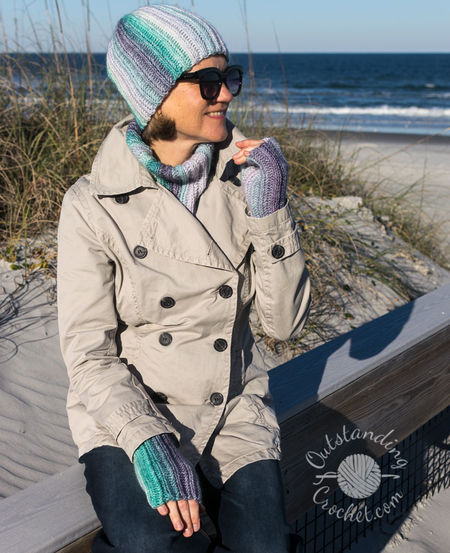 A woman wearing a coordinated Ryft crochet set in shades of green and purple. She is pictured from the chest up, sporting a chunky ribbed beanie pulled low, a thick matching cowl, and fingerless gloves. The Aran weight cotton yarn shows a seamless transition between colors, following the vertical texture of the ribs.