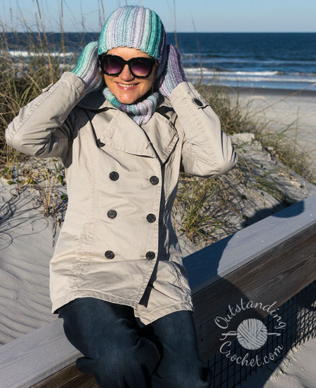 A woman wearing a coordinated Ryft crochet set in shades of green and purple. She is pictured from the chest up, sporting a chunky ribbed beanie pulled low, a thick matching cowl, and fingerless gloves. The Aran weight cotton yarn shows a seamless transition between colors, following the vertical texture of the ribs.