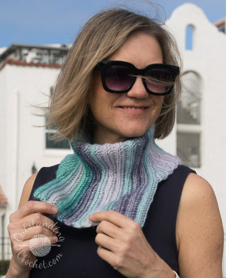 A woman wearing Ryft Crochet Neck Warmer in shades of green and purple. She is pictured from the chest up, sporting a chunky cowl. The Aran weight cotton yarn shows a seamless transition between colors, following the vertical texture of the ribs.