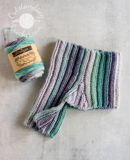 A flat lay of a unisex cowl neck warmer. The multicolored yarn creates a seamless transition between vertical stripes, highlighting the thick, vertical ribbing texture of the Aran weight cotton yarn in green and purple shades.