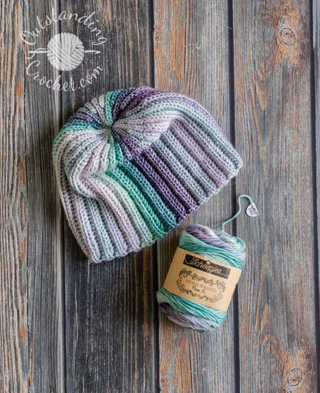 A flat lay of a unisex hat. The multicolored yarn creates a seamless transition between vertical stripes, highlighting the thick, vertical ribbing texture of the Aran weight cotton yarn in green and purple shades.