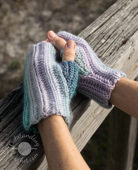Pictures shows hands in Ryft unisex fingerless gloves. The multicolored yarn creates a seamless transition between vertical stripes, highlighting the thick, vertical ribbing texture of the Aran weight cotton yarn in green and purple shades.