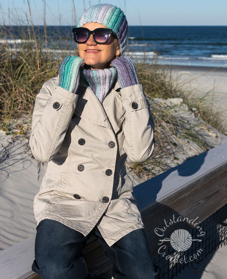 A woman wearing a coordinated Ryft crochet set in shades of green and purple. She is pictured from the chest up, sporting a chunky ribbed beanie pulled low, a thick matching cowl, and fingerless gloves. The Aran weight cotton yarn shows a seamless transition between colors, following the vertical texture of the ribs.