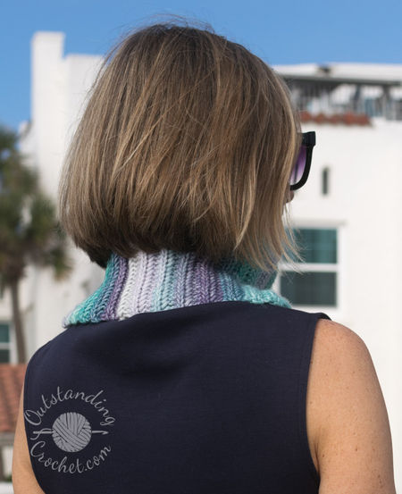 A woman wearing Ryft Crochet Neck Warmer in shades of green and purple. She is pictured from the chest up, sporting a chunky cowl. The Aran weight cotton yarn shows a seamless transition between colors, following the vertical texture of the ribs.