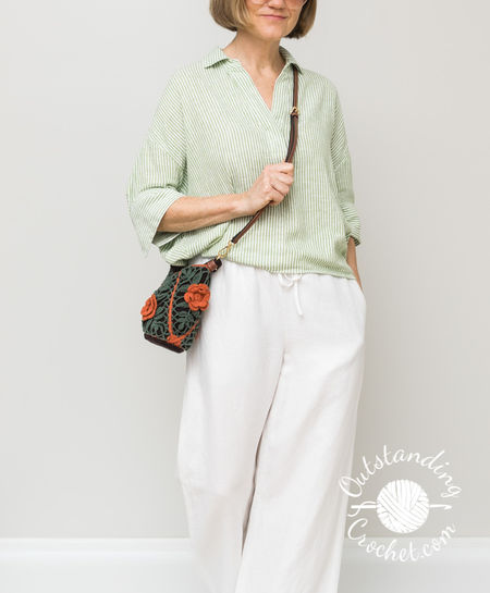 A woman wears a small crochet cross-body purse composed of 4 joined-as-you-go triangular motifs. The design features sage green leaf lace patterns over a dark brown lining, trimmed with bright orange diagonal seams and a matching 3D crochet flower on the front. Measures 20 cm wide by 17 cm high with a dark brown oval base, handle and and leather cross-body strap.