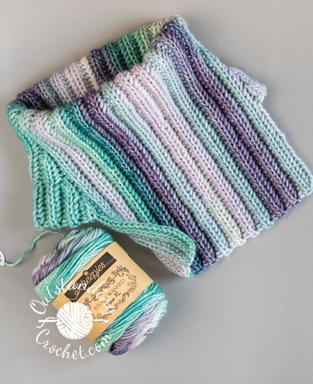 A flat lay of a Ryft unisex cowl neck warmer. The multicolored yarn creates a seamless transition between vertical stripes, highlighting the thick, vertical ribbing texture of the Aran weight cotton yarn in green and purple shades.