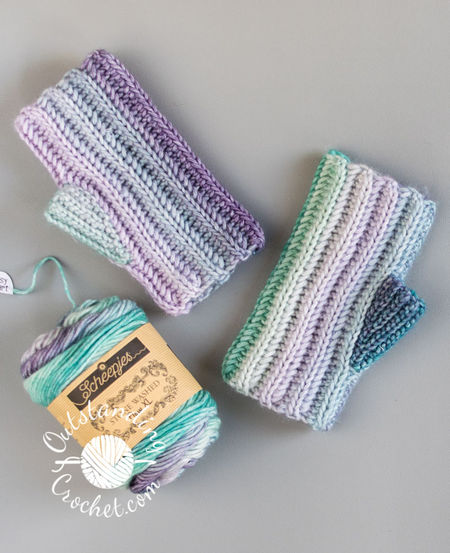 A flat lay of a Ryft unisex fingerless gloves. The multicolored yarn creates a seamless transition between vertical stripes, highlighting the thick, vertical ribbing texture of the Aran weight cotton yarn in green and purple shades.