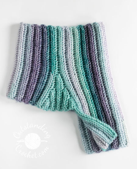 A flat lay of a unisex cowl neck warmer. The multicolored yarn creates a seamless transition between vertical stripes, highlighting the thick, vertical ribbing texture of the Aran weight cotton yarn in green and purple shades.