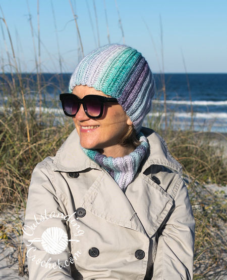 A woman wearing a coordinated Ryft crochet set in shades of green and purple. She is pictured from the chest up, sporting a chunky ribbed beanie pulled low, a thick matching cowl, and fingerless gloves. The Aran weight cotton yarn shows a seamless transition between colors, following the vertical texture of the ribs.