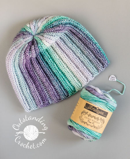 A flat lay of a unisex hat. The multicolored yarn creates a seamless transition between vertical stripes, highlighting the thick, vertical ribbing texture of the Aran weight cotton yarn in green and purple shades.