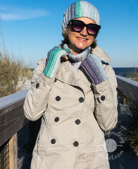 A woman wearing a coordinated Ryft crochet set in shades of green and purple. She is pictured from the chest up, sporting a chunky ribbed beanie pulled low, a thick matching cowl, and fingerless gloves. The Aran weight cotton yarn shows a seamless transition between colors, following the vertical texture of the ribs.