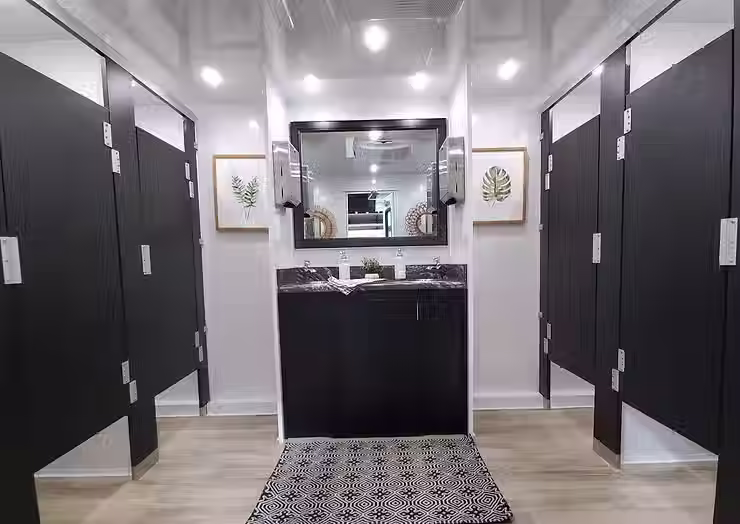 Luxury restroom trailer interior with private stalls, a central sink, large mirror, modern lighting, and wood style flooring.