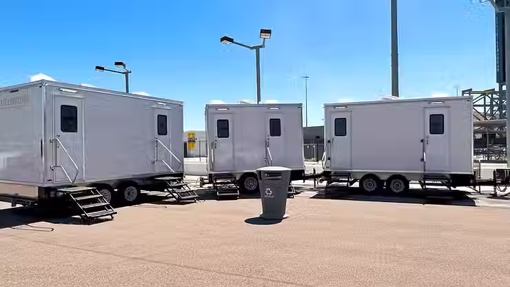Three portable restroom trailers set up in a paved outdoor area under a clear blue sky.
