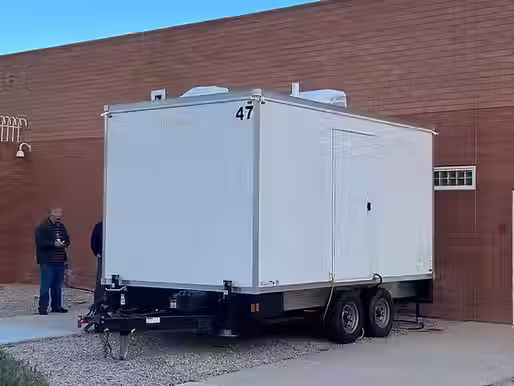 White portable restroom trailer parked beside a brick commercial building.