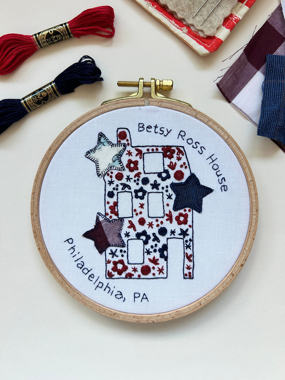 Embroidery hoop with a stitched house design labeled Betsy Ross House, surrounded by red and navy thread, fabric swatches, and needles.