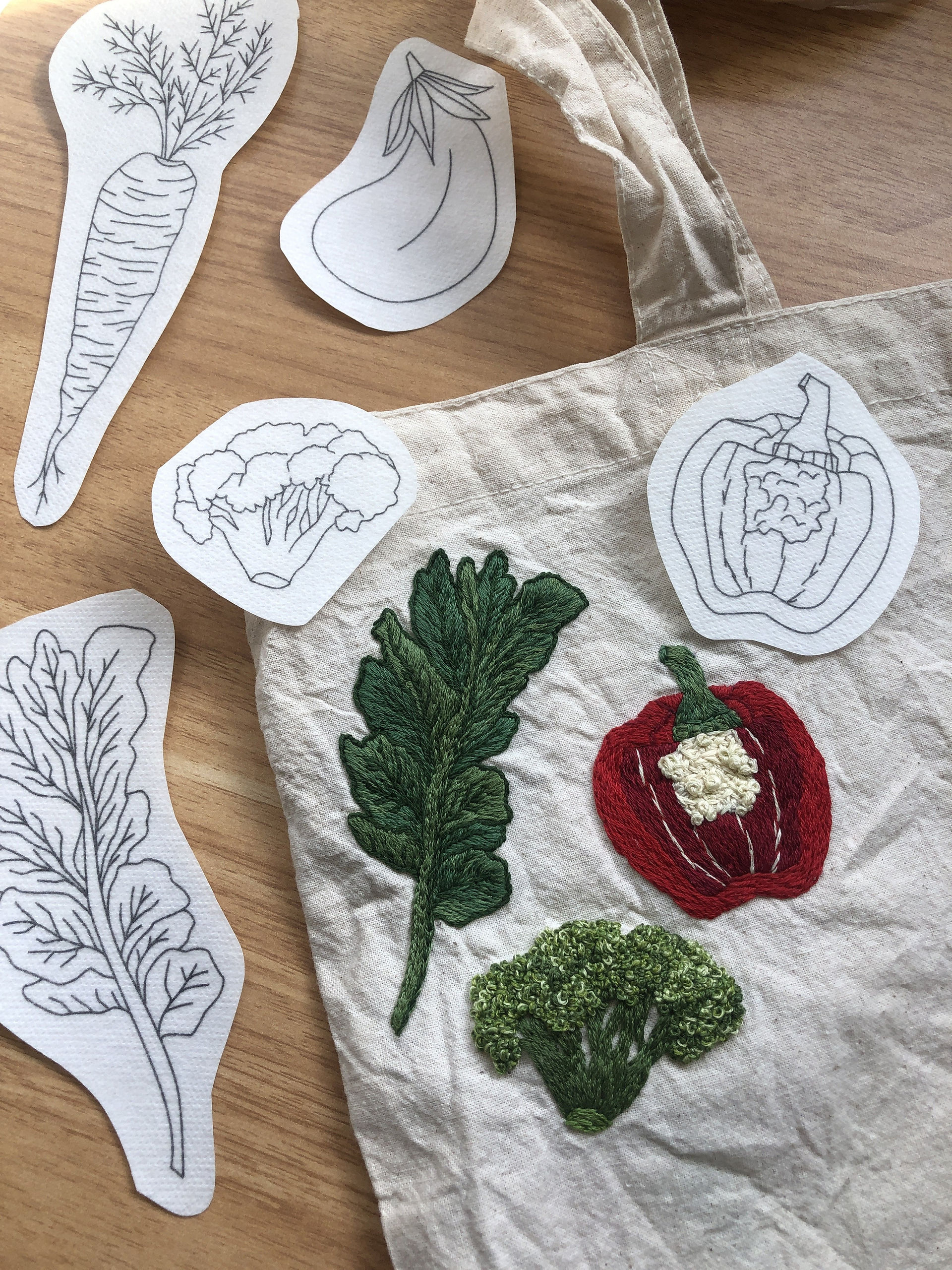 Vegetable embroidery patterns next to a tote bag embroidered with a kale leaf, red bell pepper, and broccoli. Patterns include an eggplant, carrot, kale leaf, broccoli, and pepper.