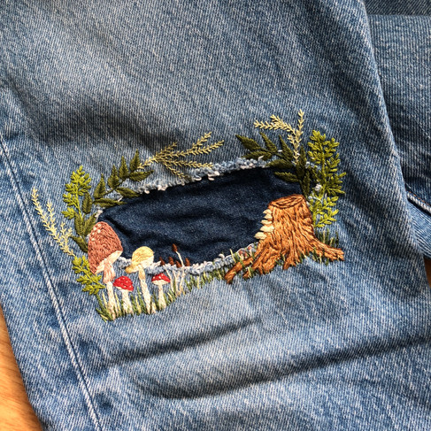 Denim jeans with a denim patch surrounded by embroidered mushrooms, a tree stump, and leafy vines.