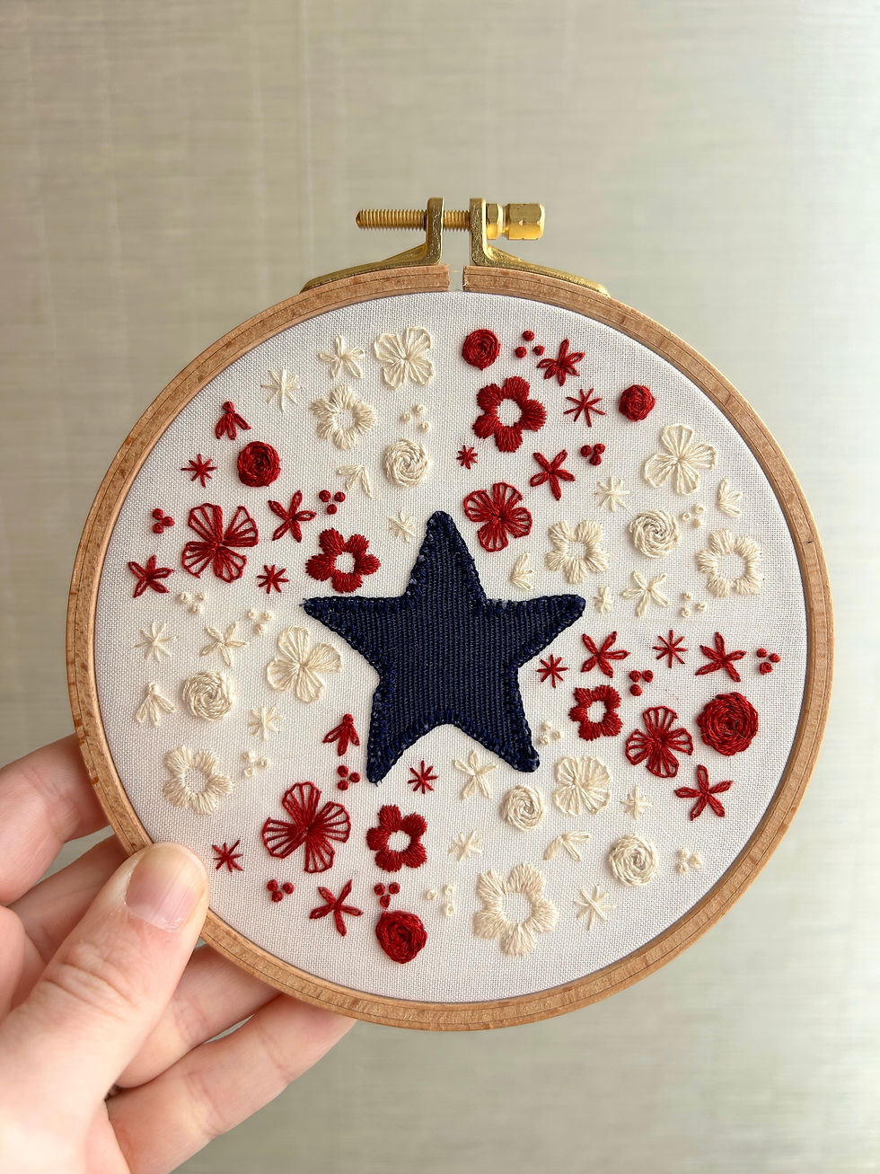 Hand holding embroidery hoop with a navy star and red-and-cream floral stitches on white fabric