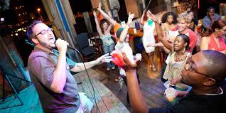 karaoke singer in a grey shit and glasses performs in front of a crowd at a small bar