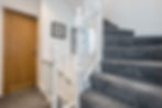 Gray carpeted staircase with white railing, wood door, and framed photos on the wall. Bright, clean interior with neutral tones.