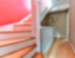 Wooden stairs with a red wall and white railing lead to a hallway with a light brown wall, a framed artwork, and a hanging bulb.