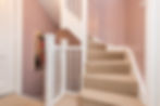 Carpeted staircase with white railing on pink walls. Art piece with text "VINS" on the wall. Beige tones create a warm ambiance.