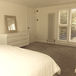 Minimalist bedroom with white bed, dresser, and mirror. Sunlit room has shutters on windows. Neutral tones create a calm, serene mood.