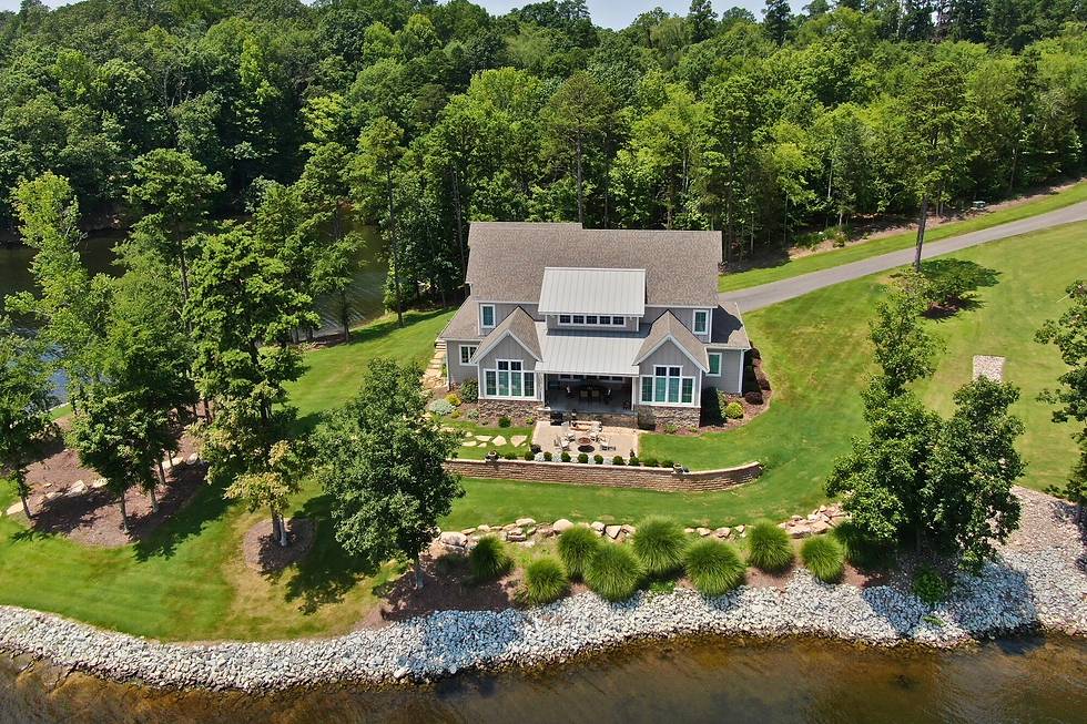 Aerial view of a large, gray house with a gabled roof by a river, surrounded by green lawn and trees, under a clear blue sky.