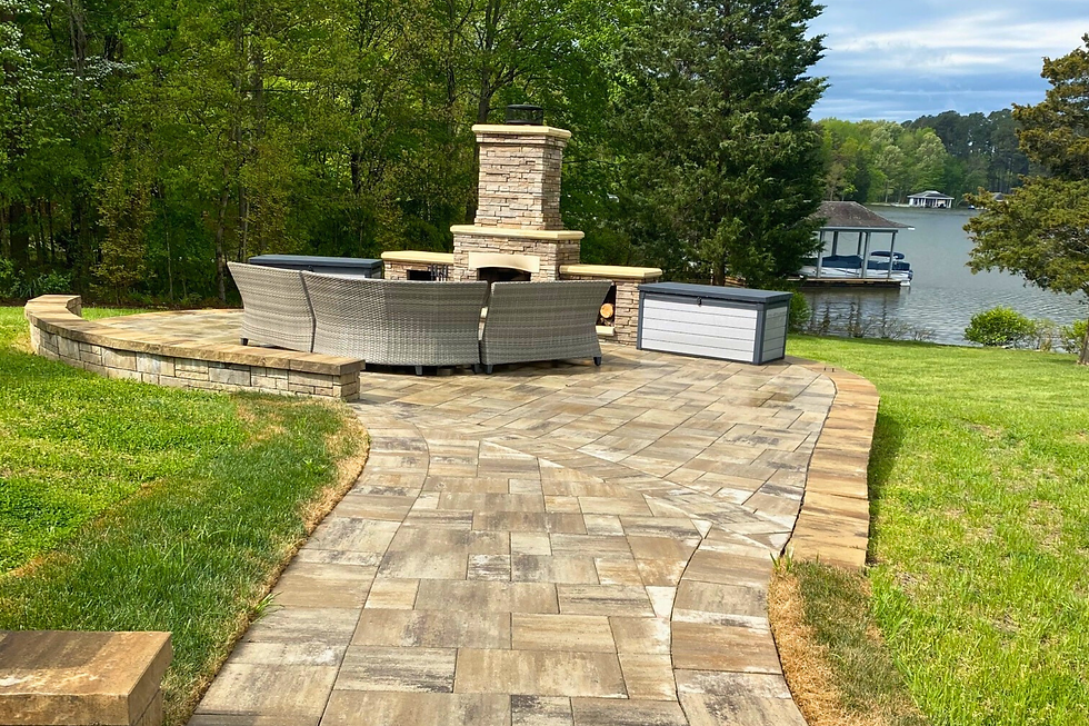 Brick patio with wicker seating and a stone fireplace by a lake. Green lawn and trees surround the area, creating a serene outdoor setting.