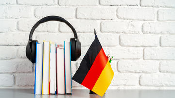 Kitapların üzerinde kulaklık bulunan bir çalışma düzeni ve yanında Almanya bayrağı; Almanca öğrenimi ve sağlık çalışanları için dil gelişimini temsil eden eğitim ortamı.