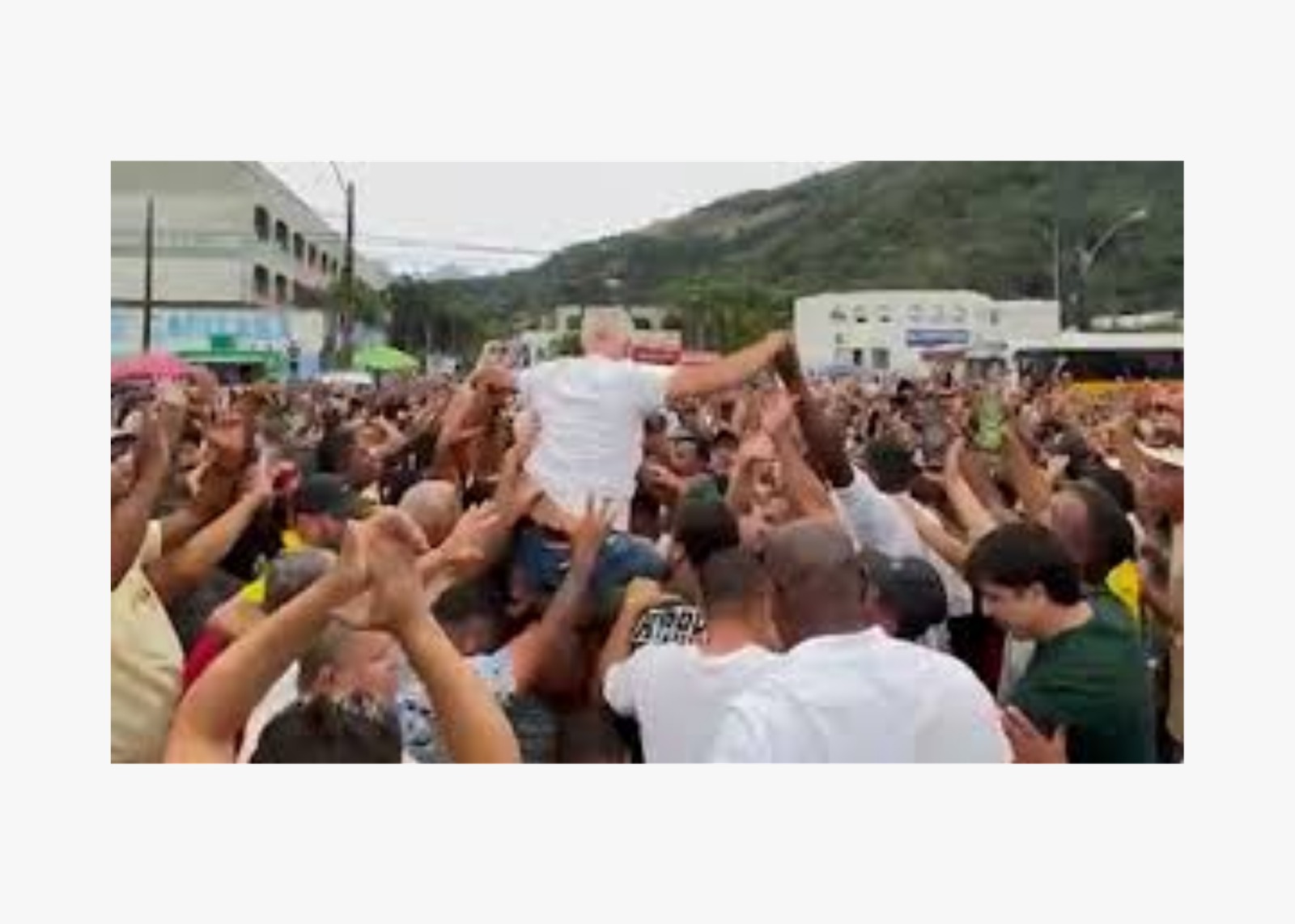 Multidão levanta uma pessoa de camiseta branca em celebração. Fundo com prédios e montanhas. Clima animado e cheio de energia.