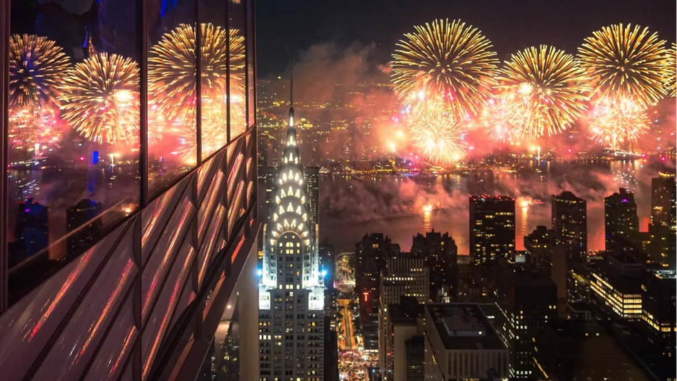 Night city skyline with fireworks lighting up the sky above waterfront. Reflections on a building's glass facade create a vibrant scene.