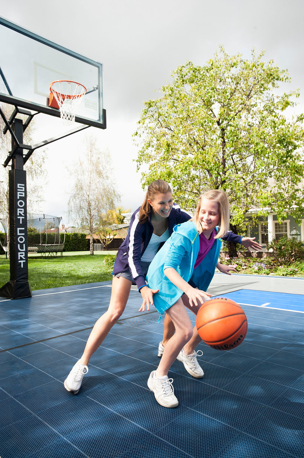 A woman and child play basketball on an outdoor court, surrounded by trees. The child, in blue, dribbles a ball while smiling.